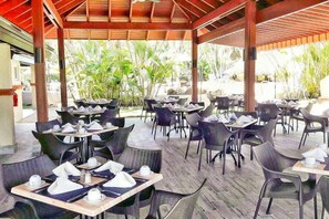 An outdoor dining area with tables and chairs, covered by a wooden roof, surrounded by greenery.