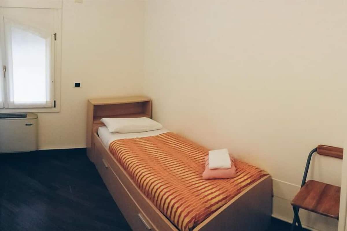 Een kleine eenpersoonskamer met een houten bedframe, een raam met zonwering en een radiator.
