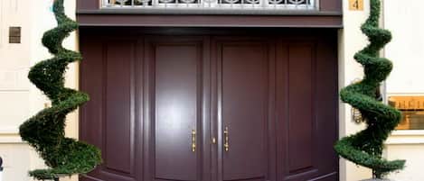 Una puerta doble de color marrón oscuro con una reja de hierro forjado en la parte superior, flanqueada por dos grandes setos topiarios.