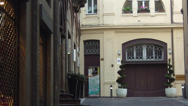 Une ruelle étroite pavée, bordée d’un immeuble doté d’une grande entrée en arc de cercle, de plantes en pot et d’une enseigne indiquant un hôtel.