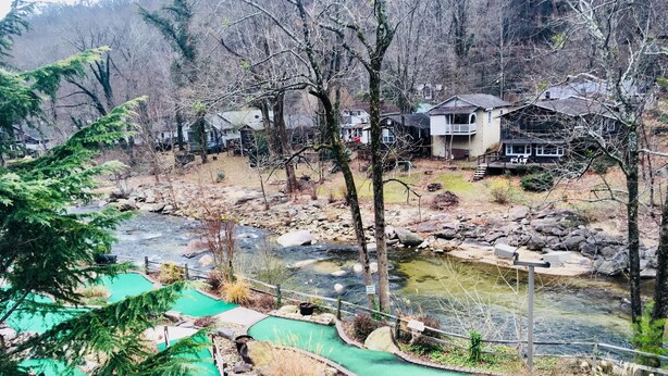 Zona junto al río con un pequeño puente, una piscina y casas al fondo.