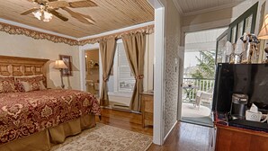 Un dormitorio con una cama grande, ventilador de techo, ventana con cortinas y balcón con mobiliario exterior.
