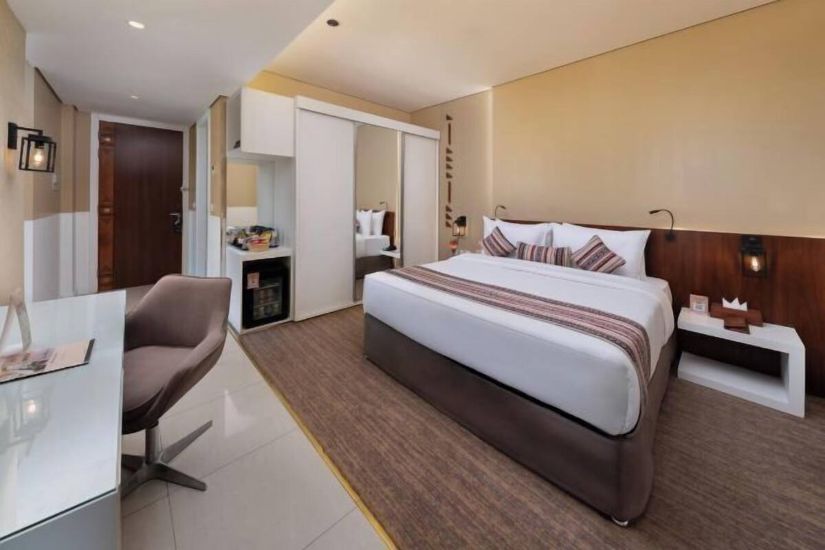A hotel room with a bed, bedside table, chair, and a desk with a lamp.