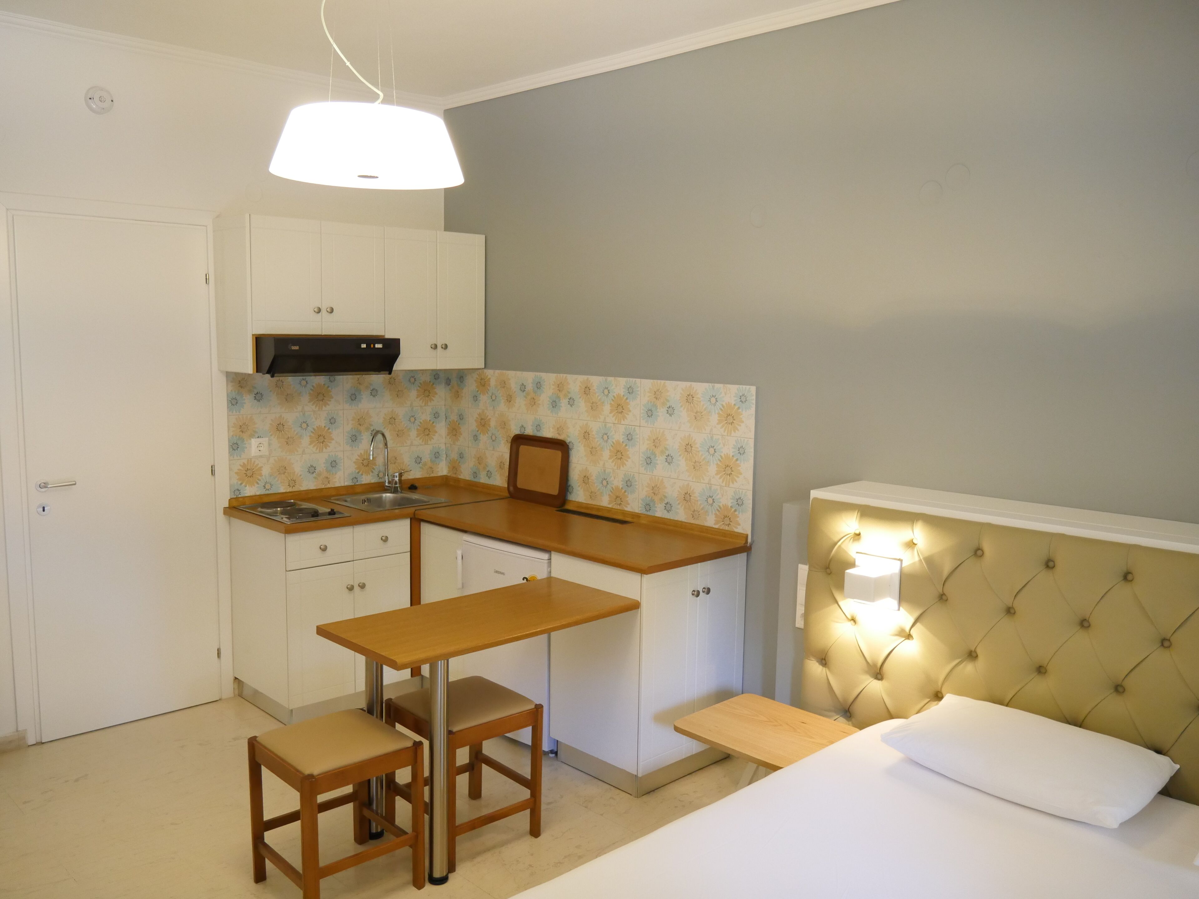 Quarto compacto com cama, mesa de cabeceira e uma pequena cozinha compacta com pia e armários.