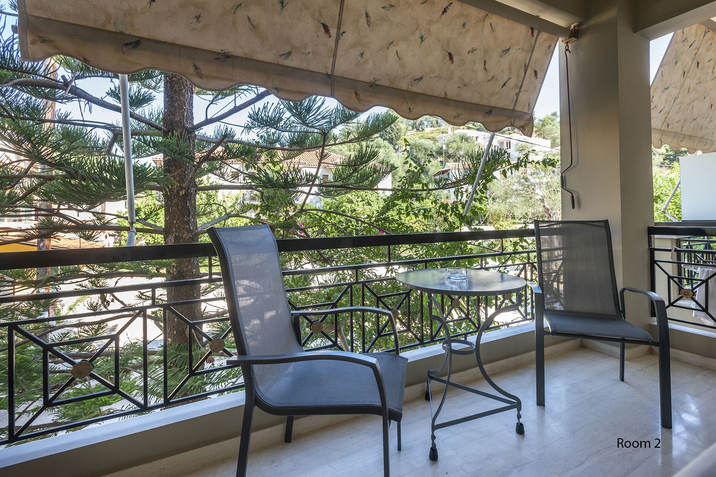 Un balcon avec deux chaises et une petite table, donnant sur un jardin avec des arbres et un bâtiment en arrière-plan.