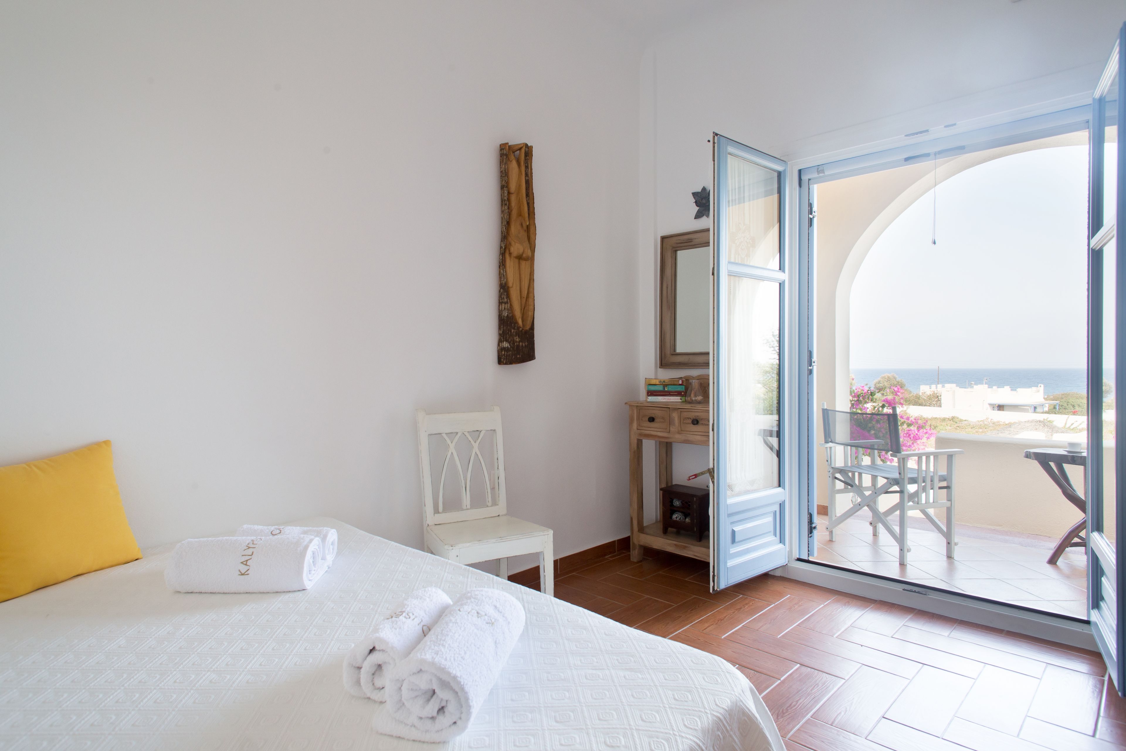 A hotel room with a bed, a chair, a small table, a mirror, and an open door leading to a balcony with a view.