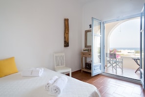 A hotel room with a bed, a chair, a small table, a mirror, and an open door leading to a balcony with a view. - Theros Houses (Santorini)