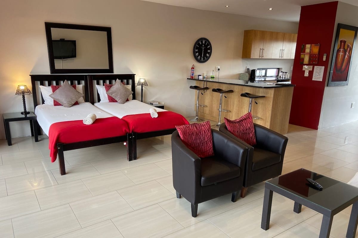 Habitación de hotel con cama, dos sillas, una mesita, televisor, reloj y kitchenette.