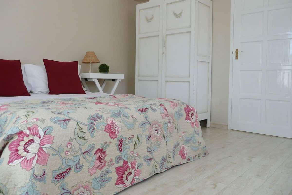 Quarto com colcha florida, guarda-roupa branco e uma pequena mesa com abajur.