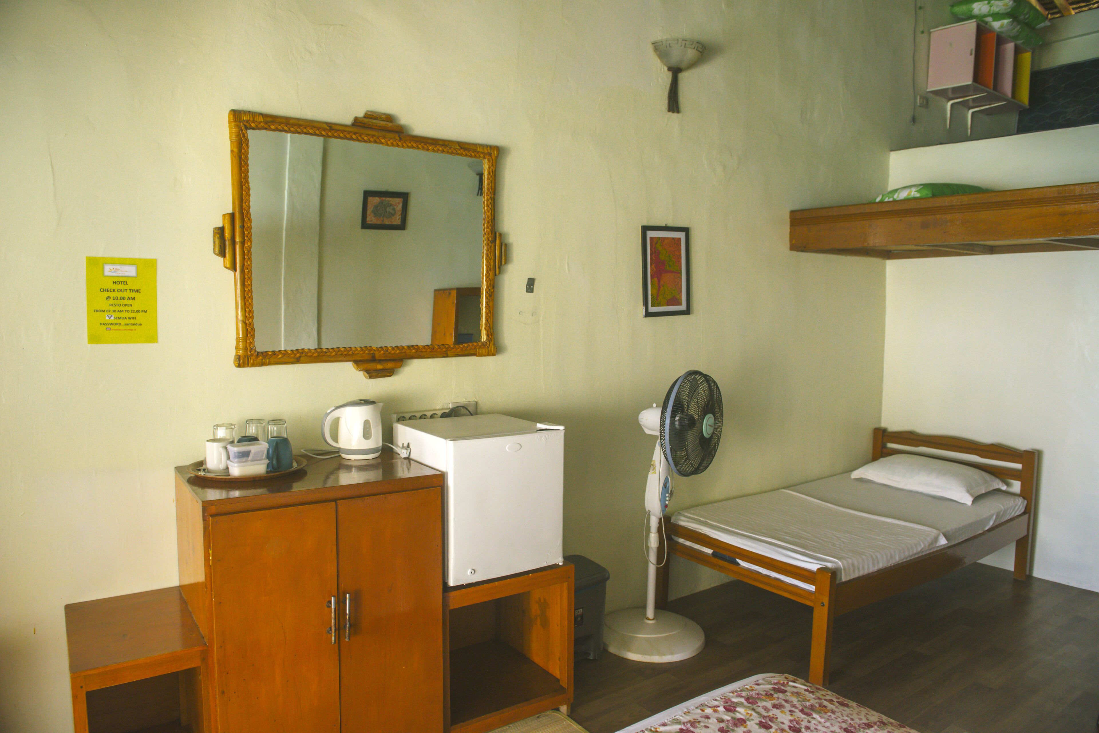 A small, simple room with a wooden bed, a small desk with a mirror, a white refrigerator, and a standing fan.