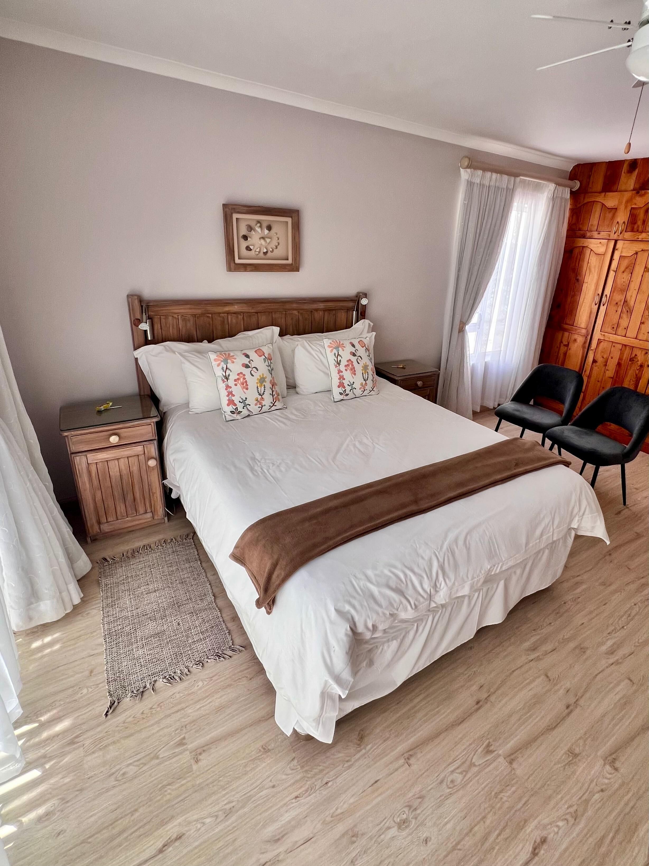 A neatly arranged bedroom with a wooden headboard, a bed with floral pillows, a bedside table with a lamp, a wooden wardrobe, and two black chairs.