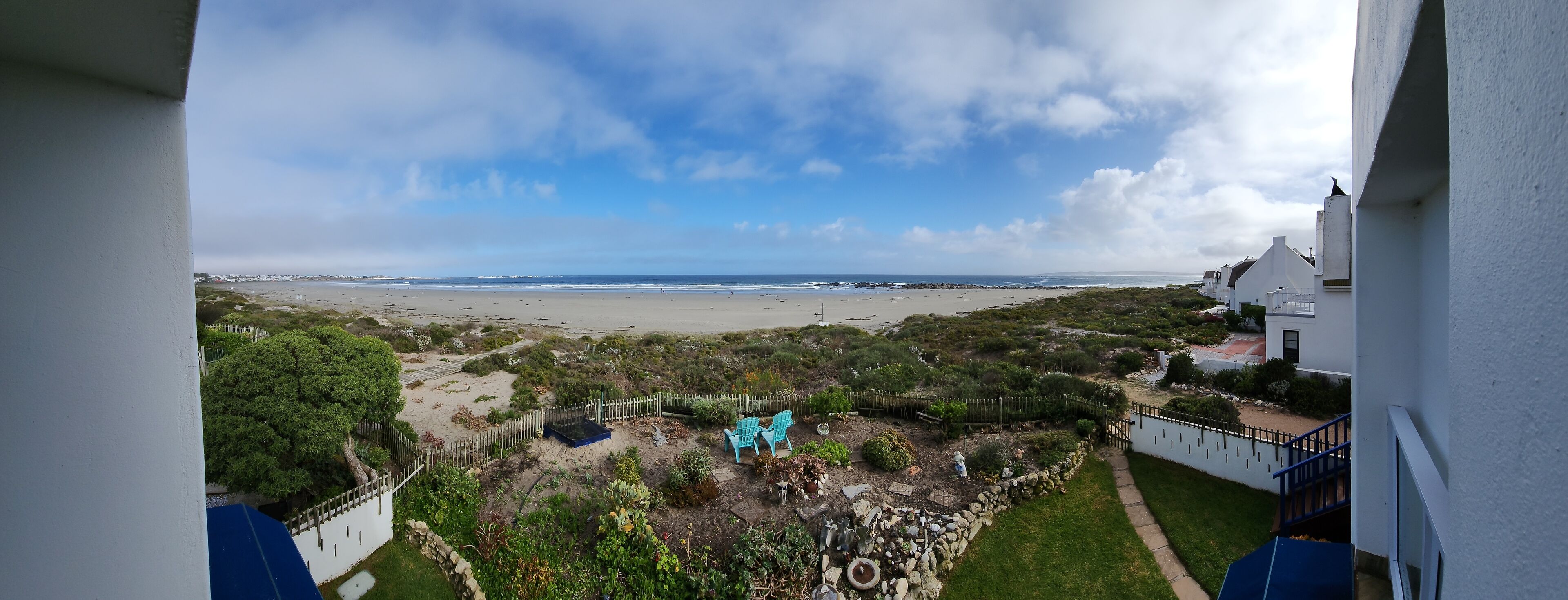 A coastal view from a balcony overlooking a beach, with a fenced garden area and a white building with a blue staircase.