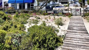 A white building with a blue awning, a wooden deck, and a sandy landscape with cacti and shrubs.