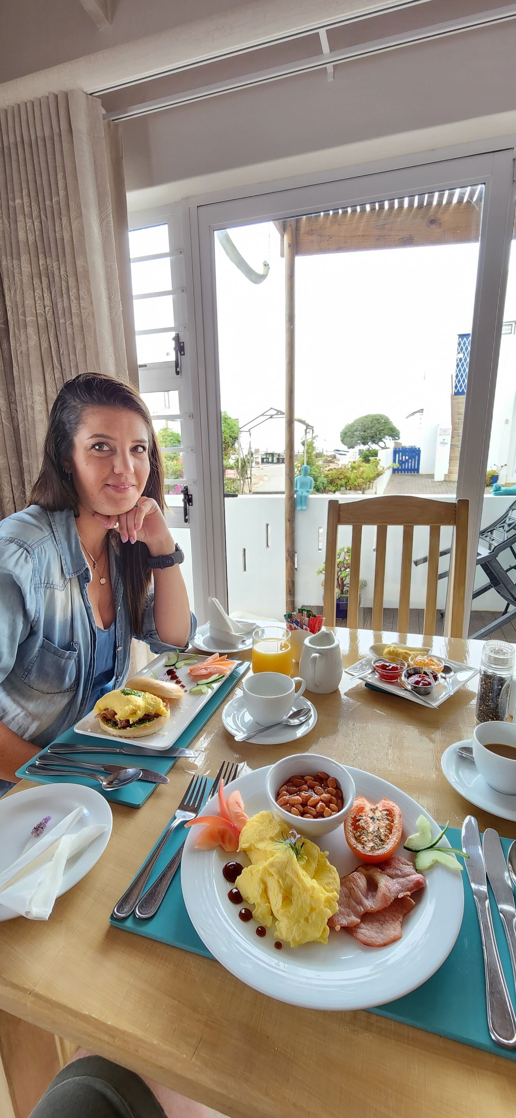 Una persona sentada a una mesa con alimentos para el desayuno, como huevos, tocino y fruta, con vista a un balcón y un espacio al aire libre.