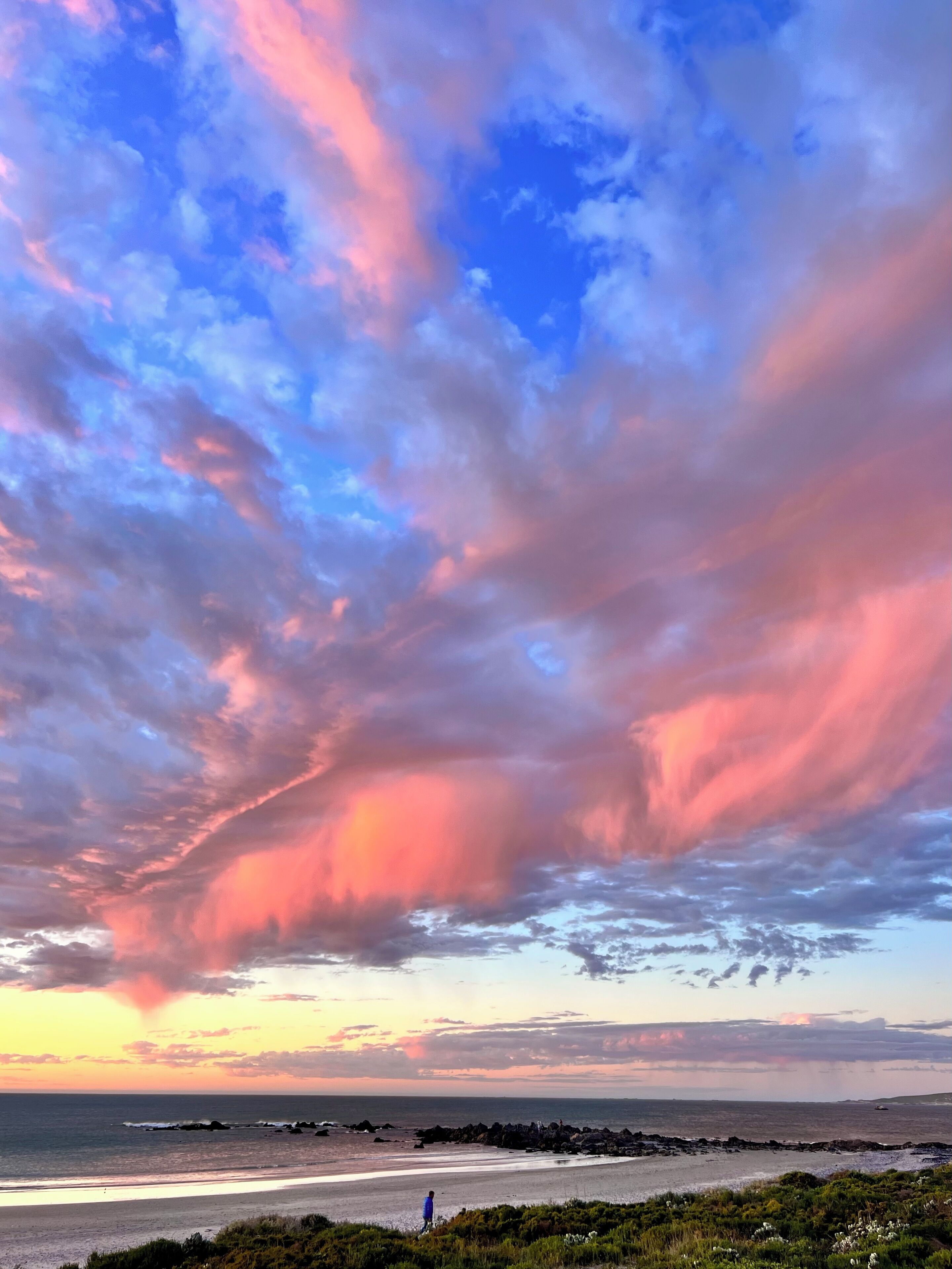 Une plage où une personne se promène, un rivage rocheux et un ciel au coucher du soleil.