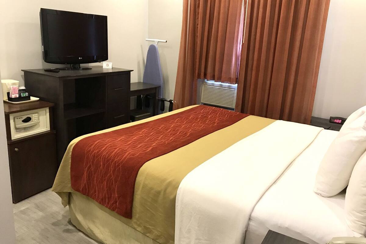Ein Hotelzimmer mit Bett, Fernseher auf einem Schrank, Schreibtisch mit Telefon und einem Fenster mit Vorhängen.