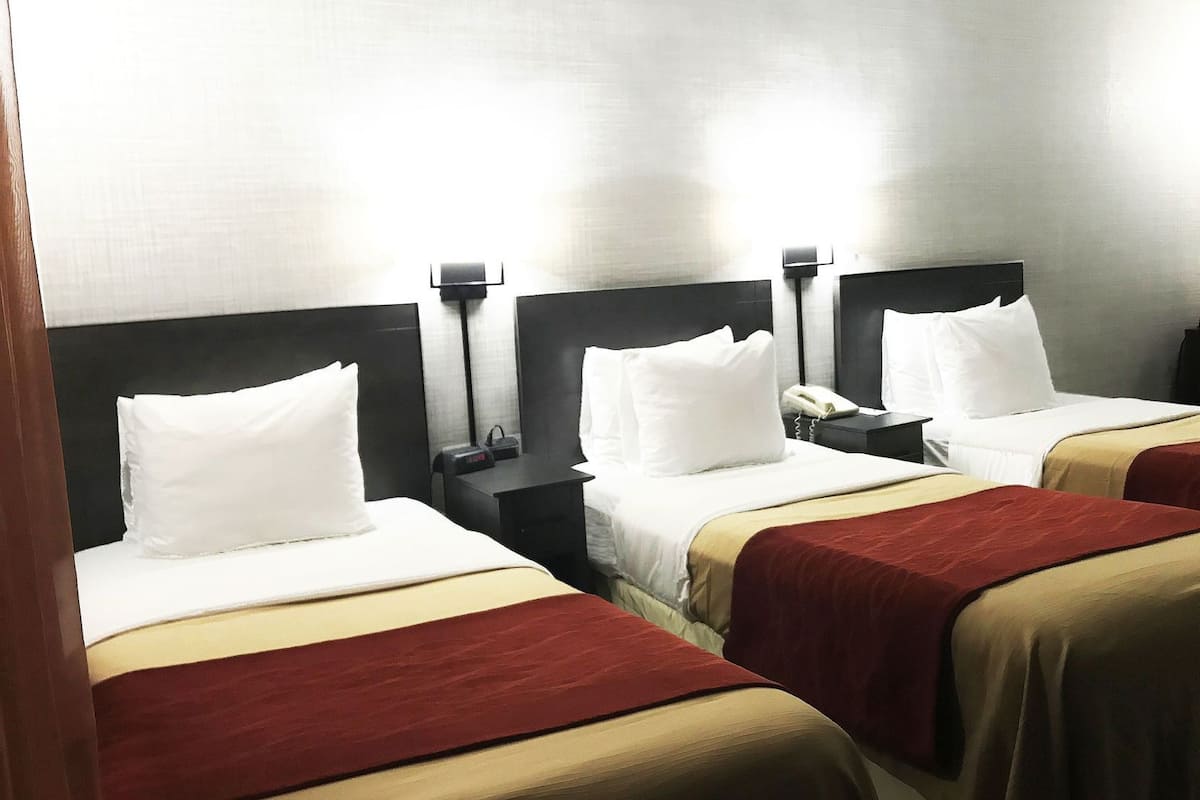 Ein Hotelzimmer mit drei Betten, jedes mit weißen Kissen und roten und beigen Bettdecken. Es gibt Nachttische mit Lampen und einem Telefon.