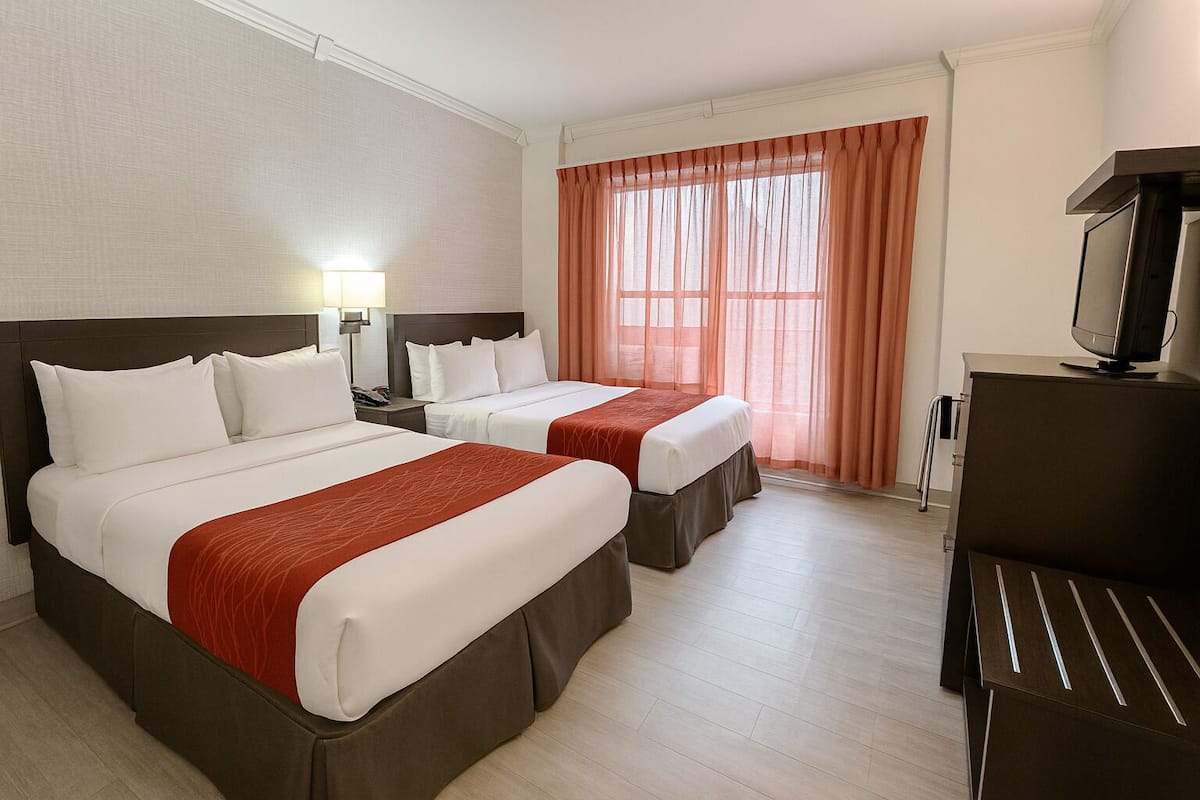 Ein Hotelzimmer mit zwei Betten, einem Fernseher, einem Schreibtisch und einem Vorhangfenster.