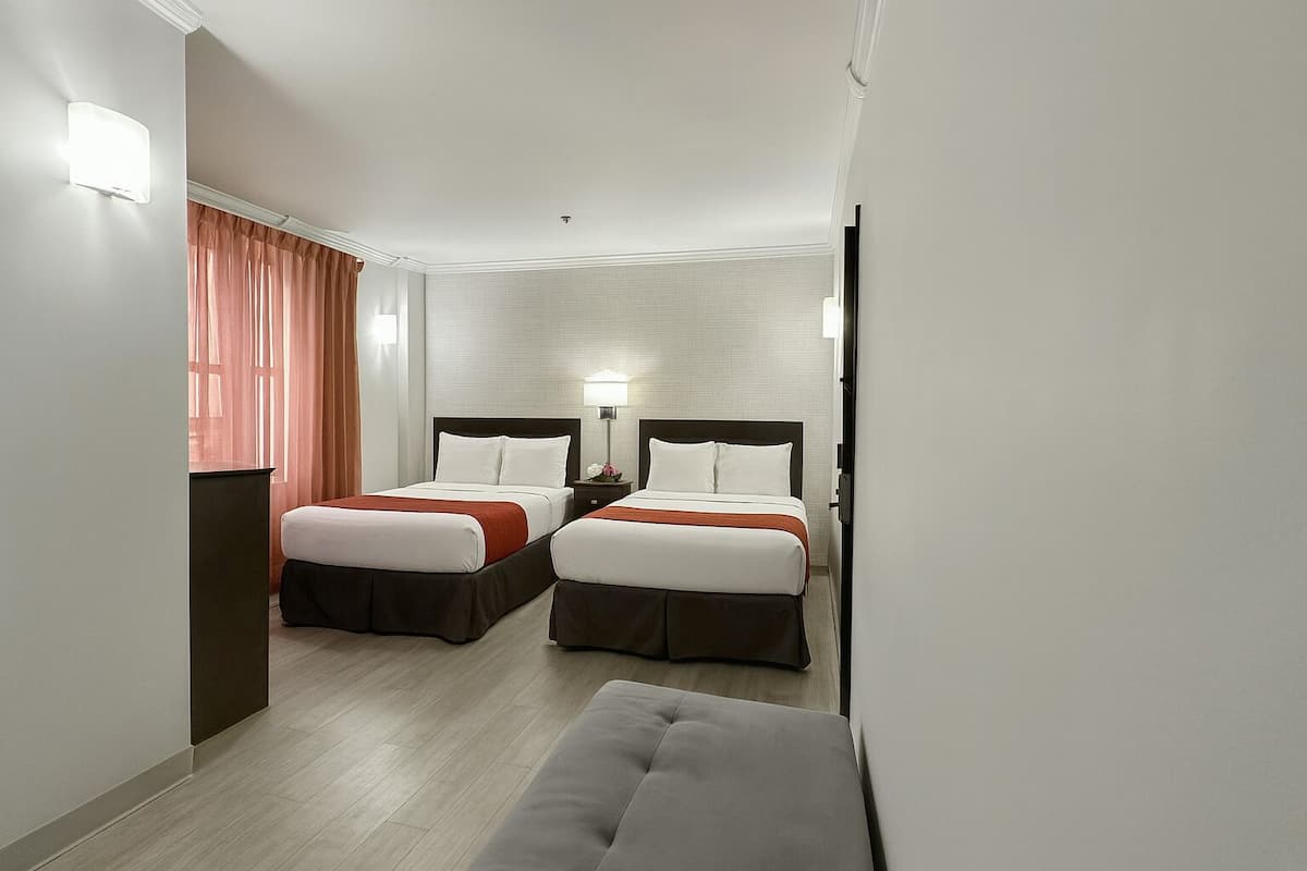 Ein Hotelzimmer mit zwei Betten, einem Nachttisch mit Lampe, einer Bank und Blick auf den Flur.