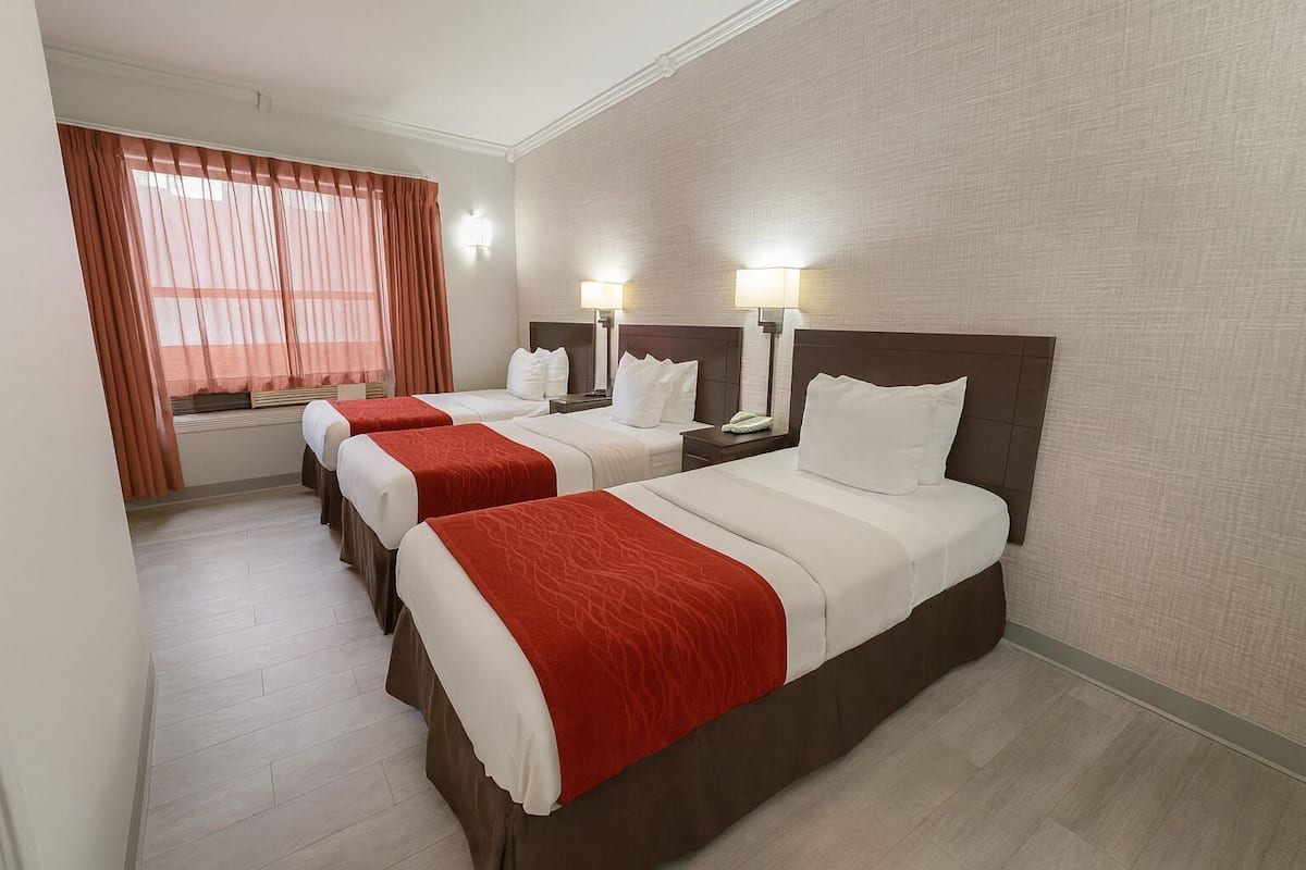 Ein Hotelzimmer mit drei Betten, jedes mit weißer Bettwäsche und roten Tagesdecken. Es gibt Nachttischlampen, ein Fenster mit Vorhängen und einen Holzboden.