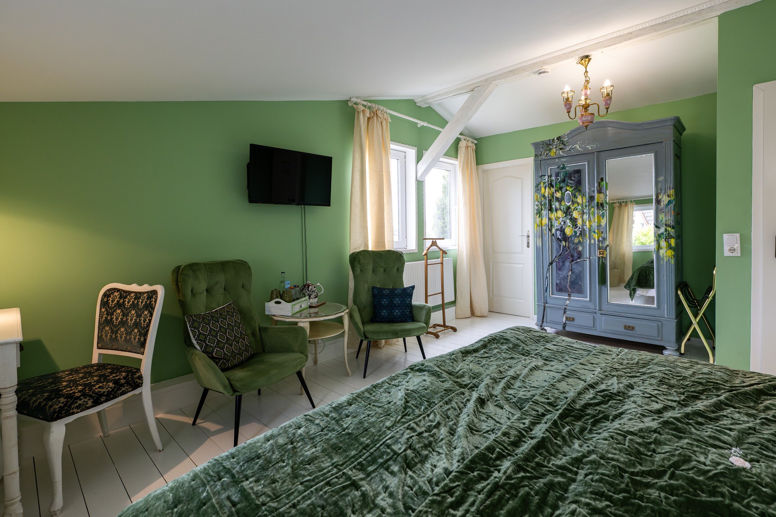 Un dormitorio con pared verde, una cama grande, dos sillones verdes, una mesita pequeña, un televisor montado en la pared y un armario con espejo.
