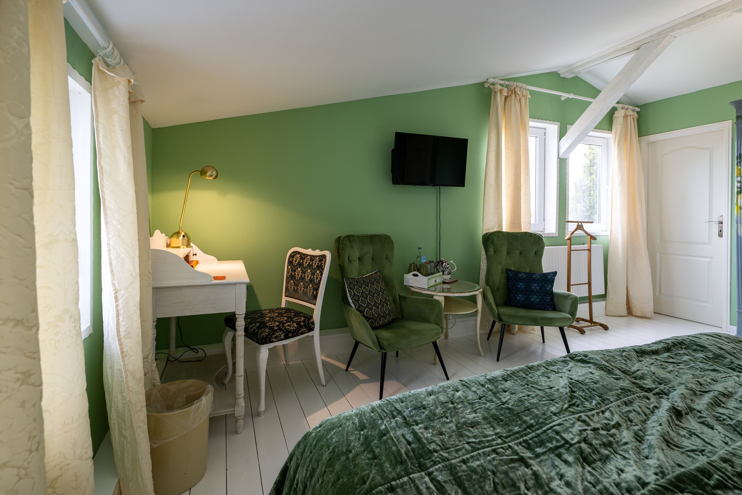 Una habitación con una cama, dos sillones verdes, una mesita con una lámpara, un televisor en la pared y un ventanal con cortinas.