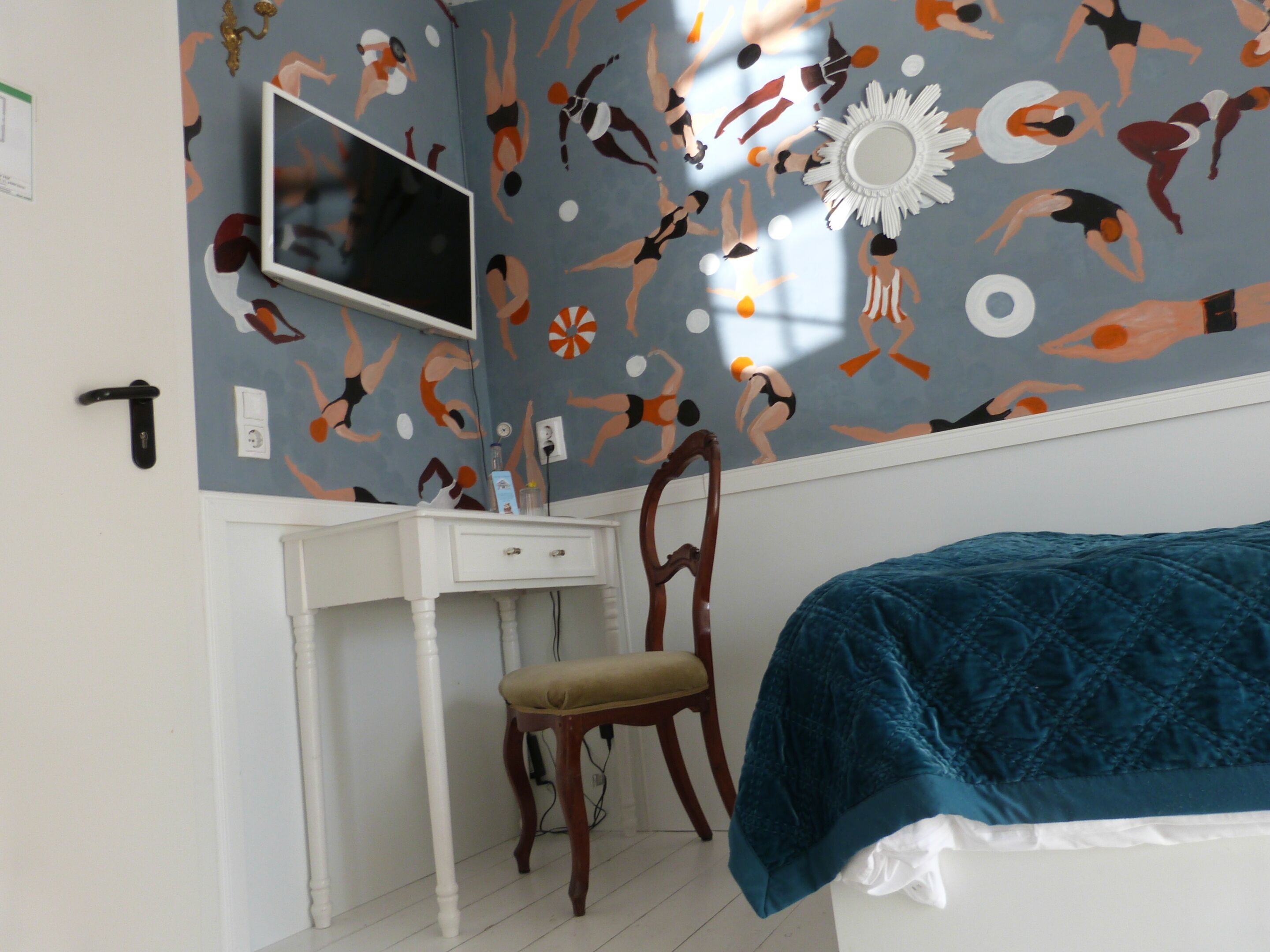 A bedroom with a bed, a desk, and a chair. The wall has a pattern of swimmers.