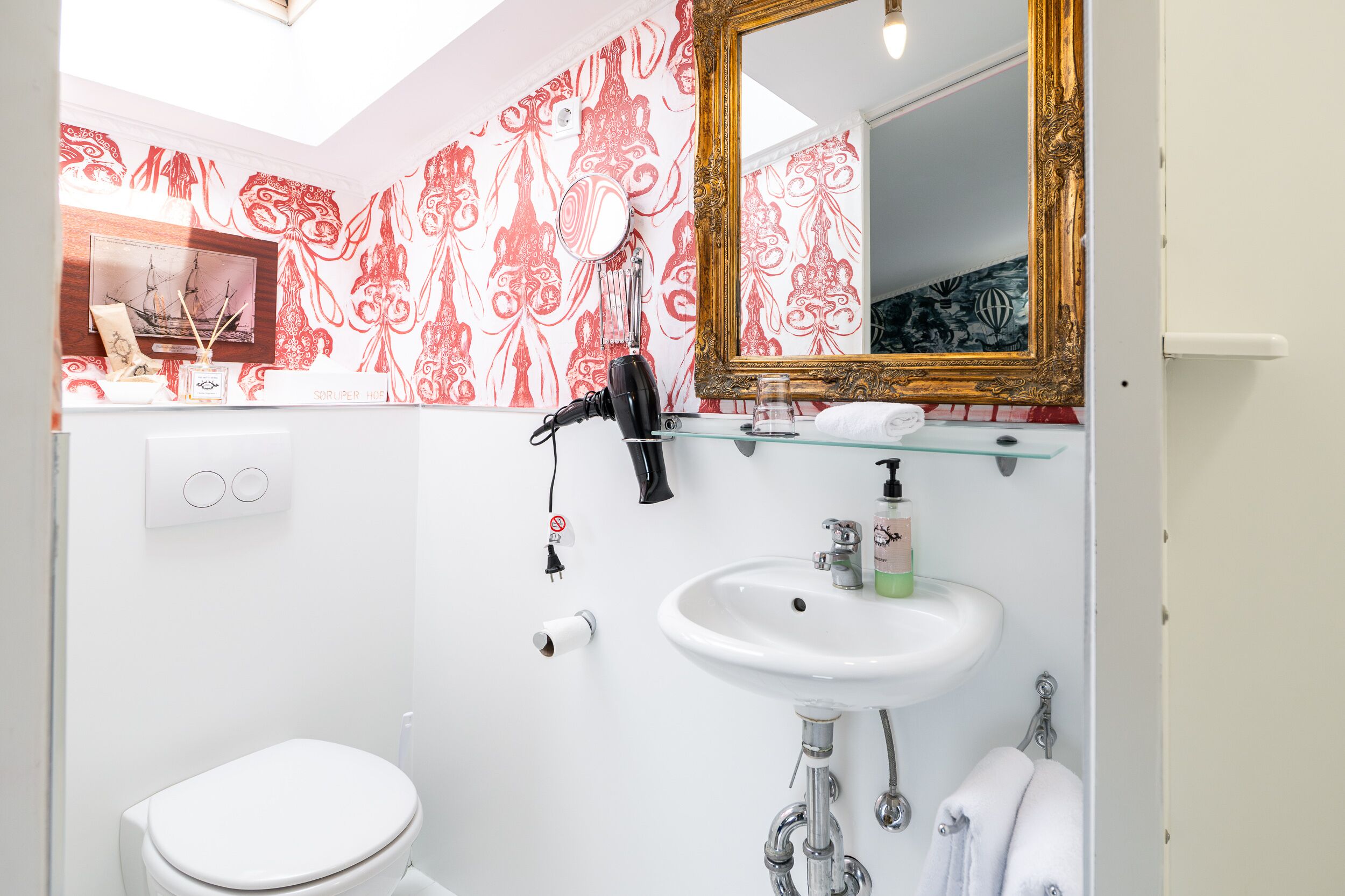 Ein Badezimmer mit einer weißen Toilette, einem Waschbecken mit einer Glasablage, einem Spiegel mit aufwendigem Rahmen und Tapete mit Muster.