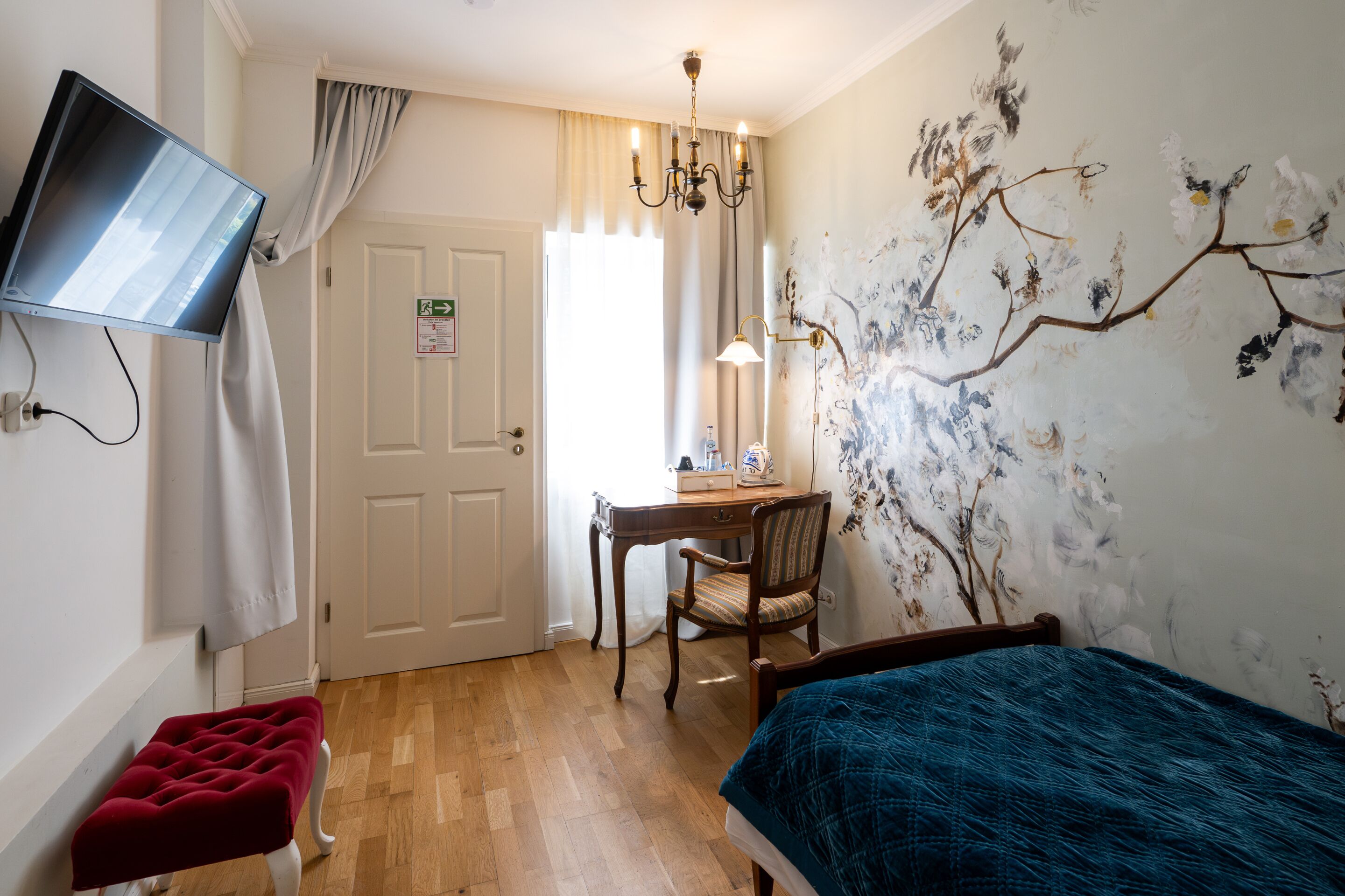 A room with a bed, a desk, a chair, a television mounted on the wall, and a painting on the wall.