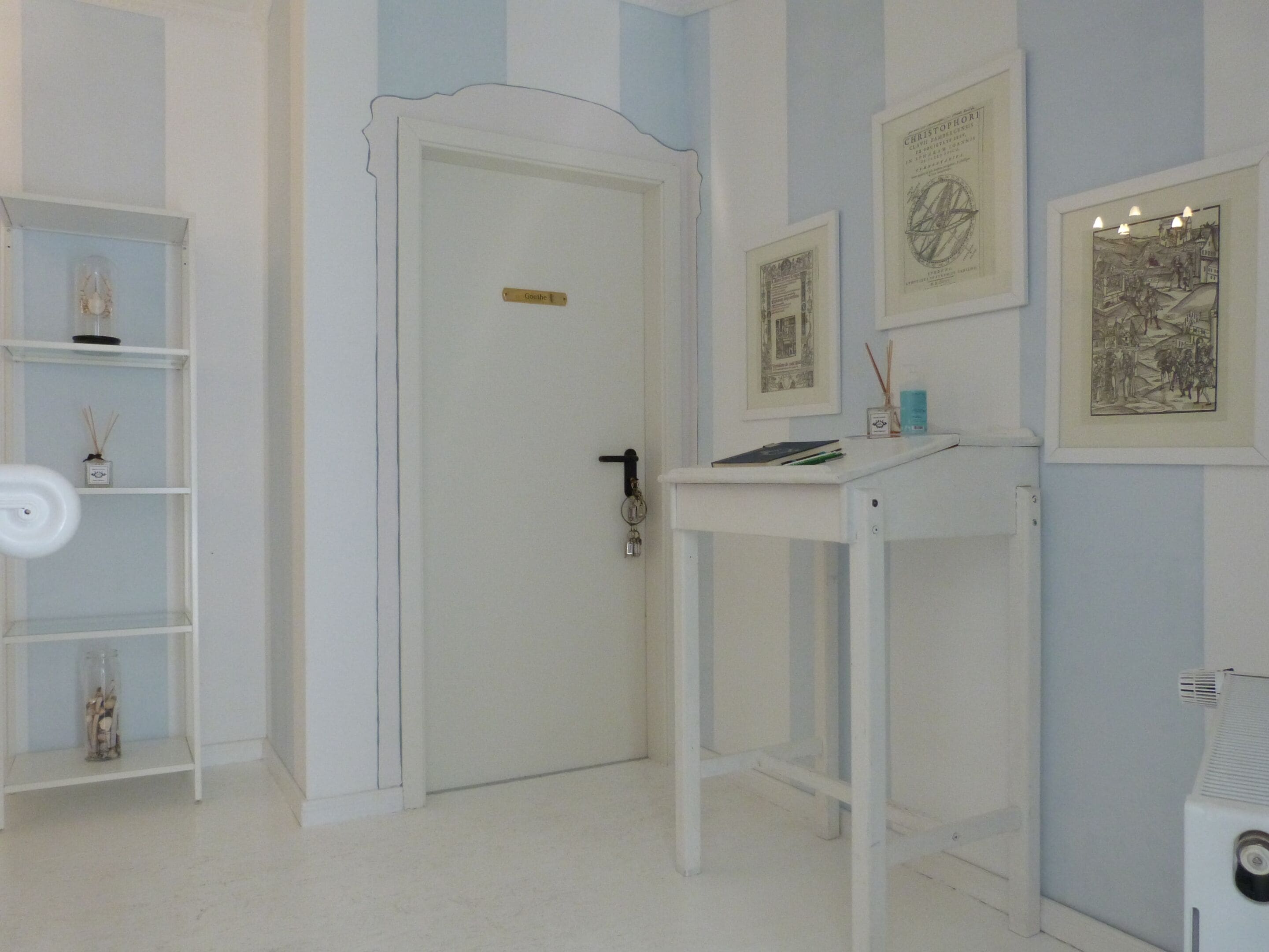Een kamer met een witte deur, een wit plankje met decoratieve objecten, een witte tafel met een boek en een klein bakje, en lijstwerk aan de muur.