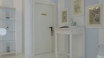 Een kamer met een witte deur, een wit plankje met decoratieve objecten, een witte tafel met een boek en een klein bakje, en lijstwerk aan de muur.