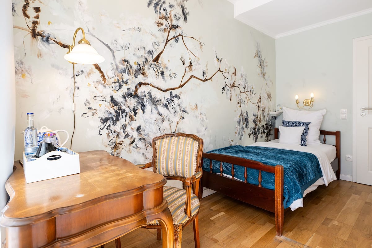 Una habitación con un escritorio de madera, una silla, una cama con manta azul y un cuadro de un árbol en la pared.