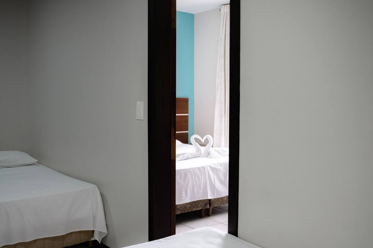 Una habitación con dos camas individuales, un cabecero de madera y una puerta que da a otra habitación con una cama y una cortina.