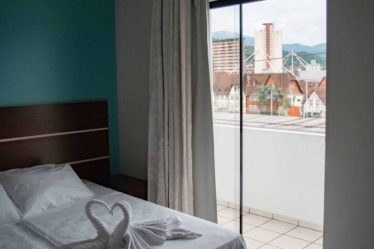Una habitación de hotel con una cama, vistas de la ciudad a través de un ventanal grande y una cortina.