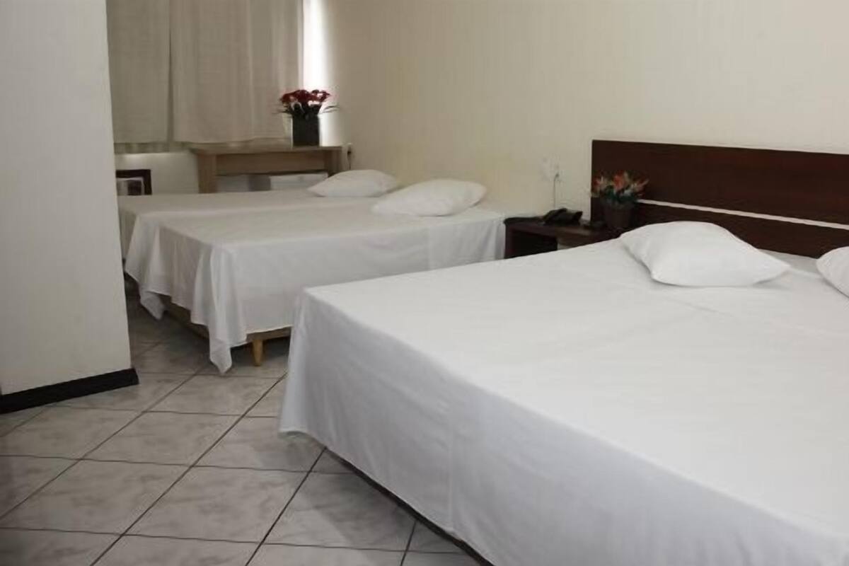 Habitación de hotel con dos camas individuales, cabecera de madera, mesita de noche con lámpara y ventana con cortinas.