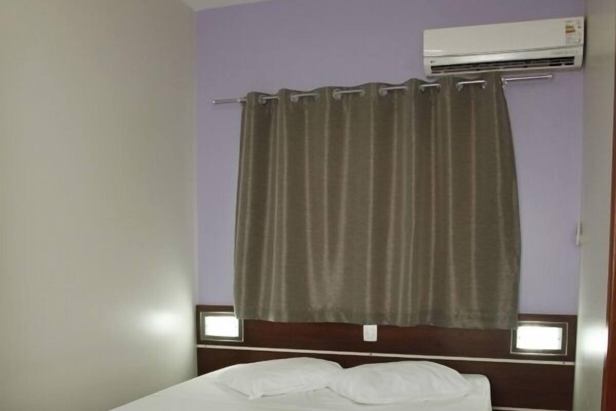 Una cama individual con cubrecama blanco en una habitación con paredes moradas, una cortina marrón y un equipo de aire acondicionado sobre la cama.