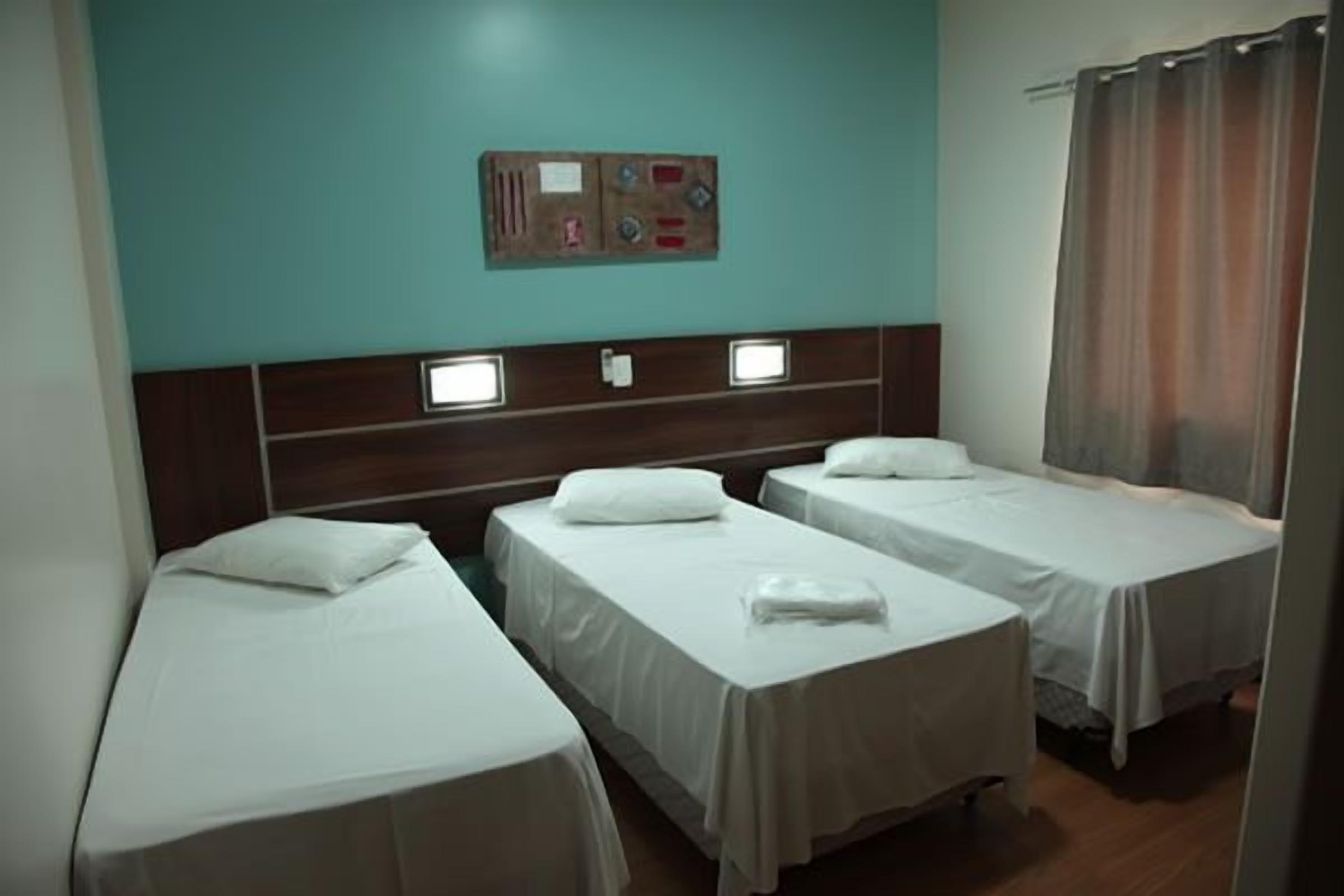 A hotel room with three single beds, a control panel on the wall, and a curtain on the window.
