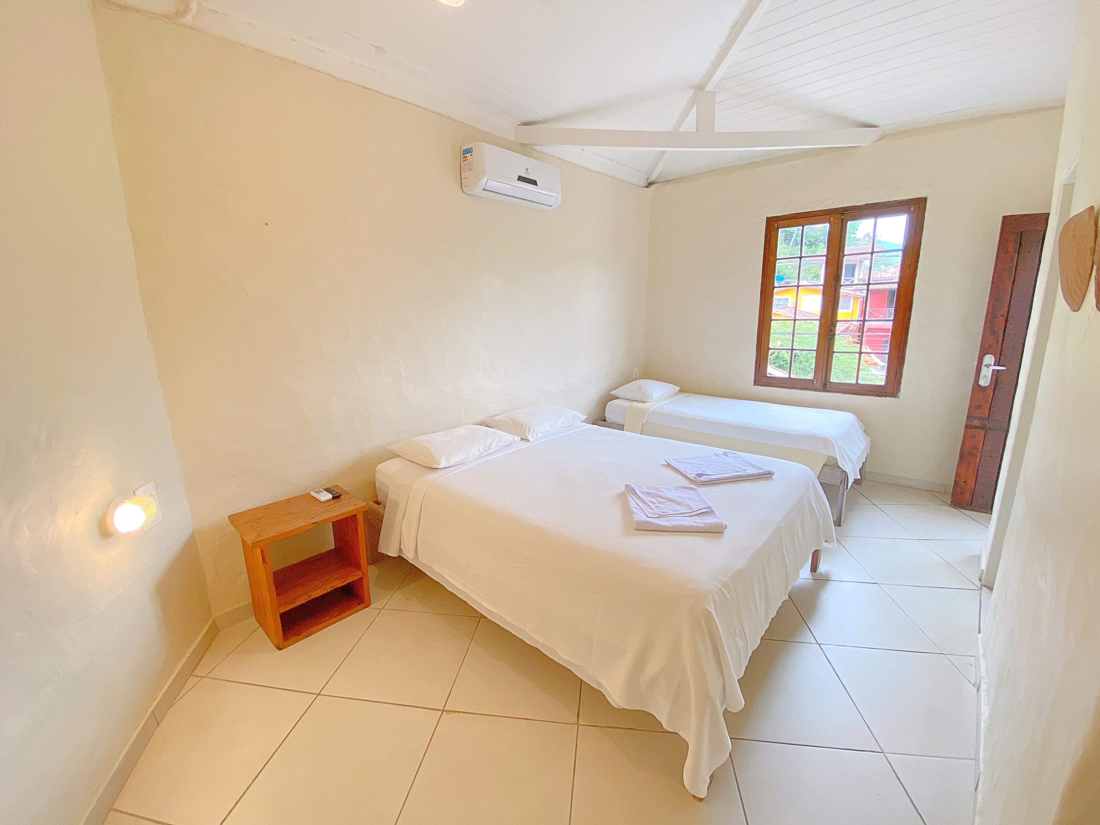 A clean, well-lit room with two beds, a small wooden nightstand, a window with a view, and an air conditioning unit.