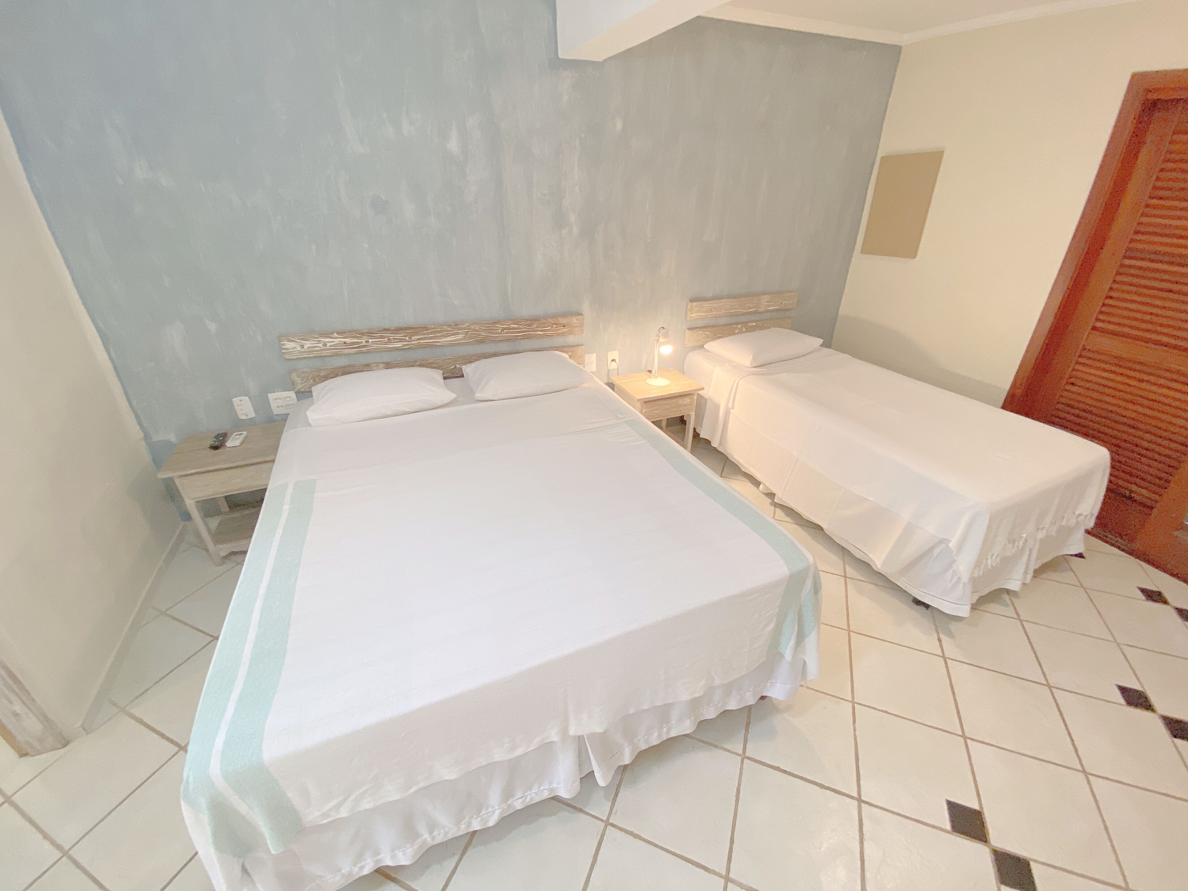 Une chambre d’hôtel avec deux lits simples, une table de chevet avec une lampe, une porte en bois et un sol carrelé.
