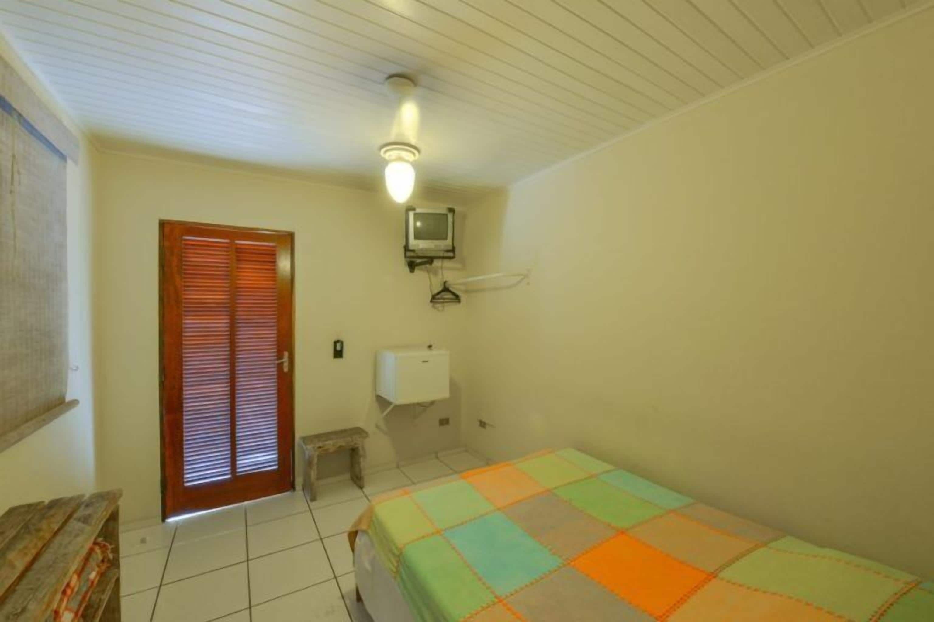 A small, simple room with a bed, a wooden bench, a toilet, and a television mounted on the wall.