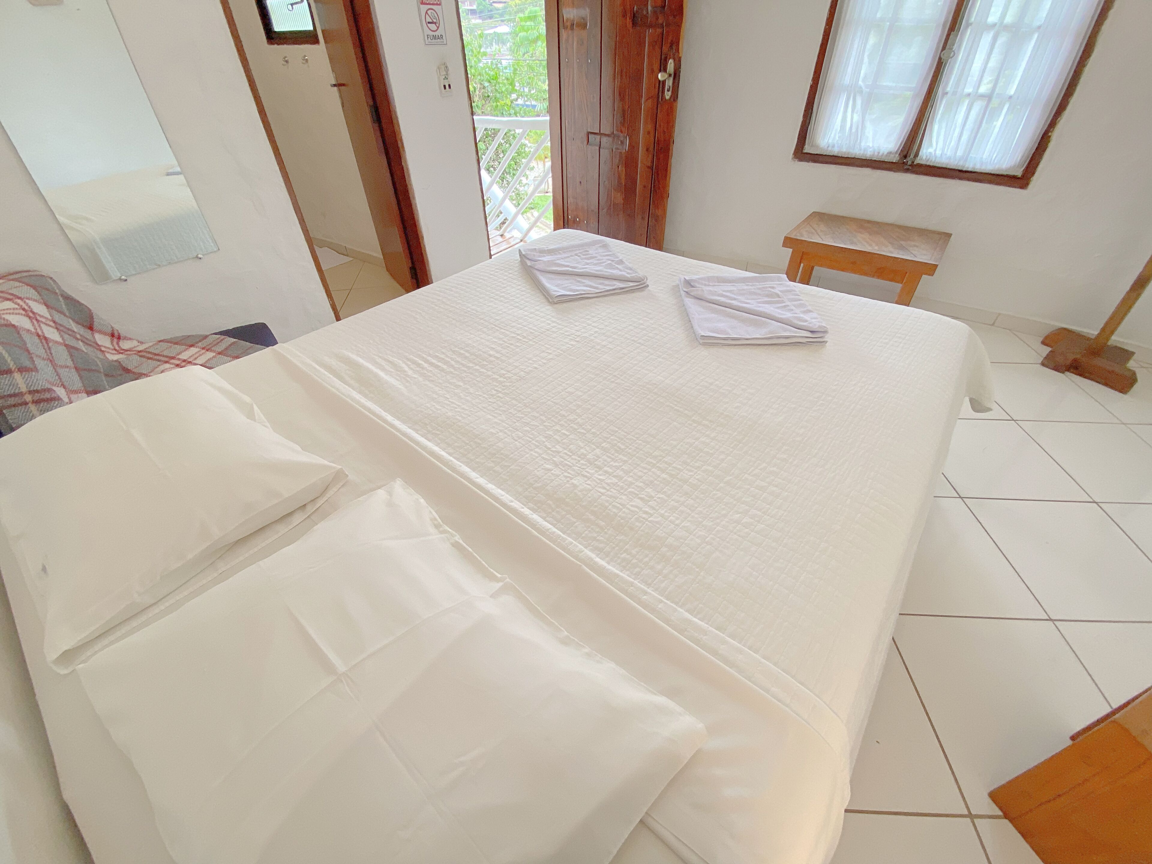 Un lit bien fait, avec du linge de lit blanc, une porte en bois sur laquelle est apposé un panneau « Interdiction de fumer », une petite table en bois et une fenêtre avec des rideaux.