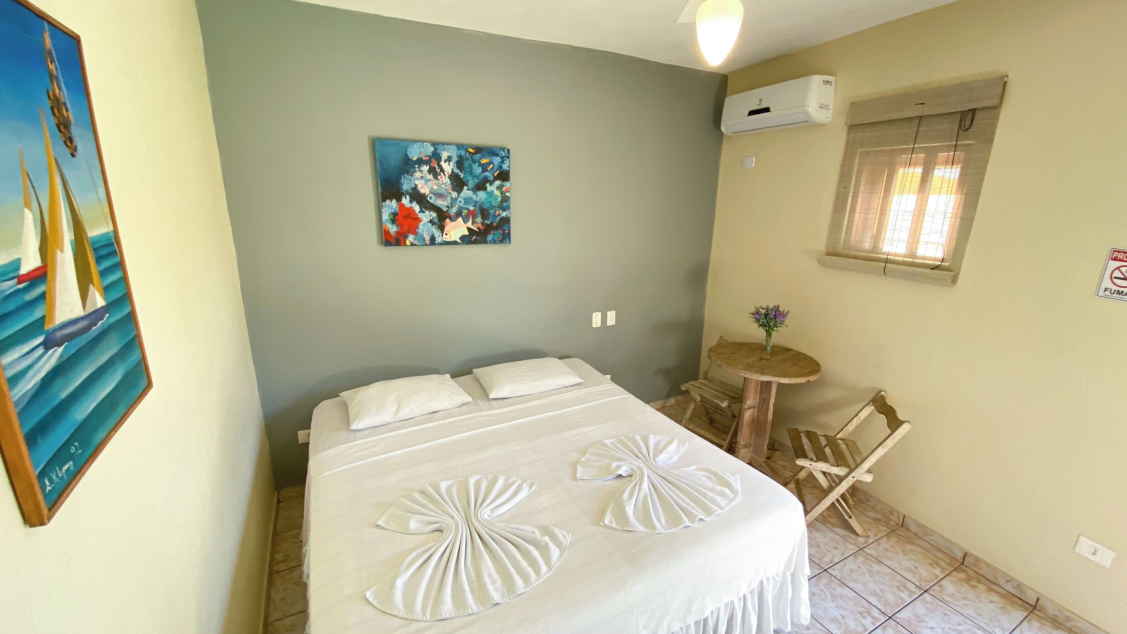 Quarto com cama, uma mesinha redonda, uma cadeira, um quadro e um aparelho de ar condicionado.
