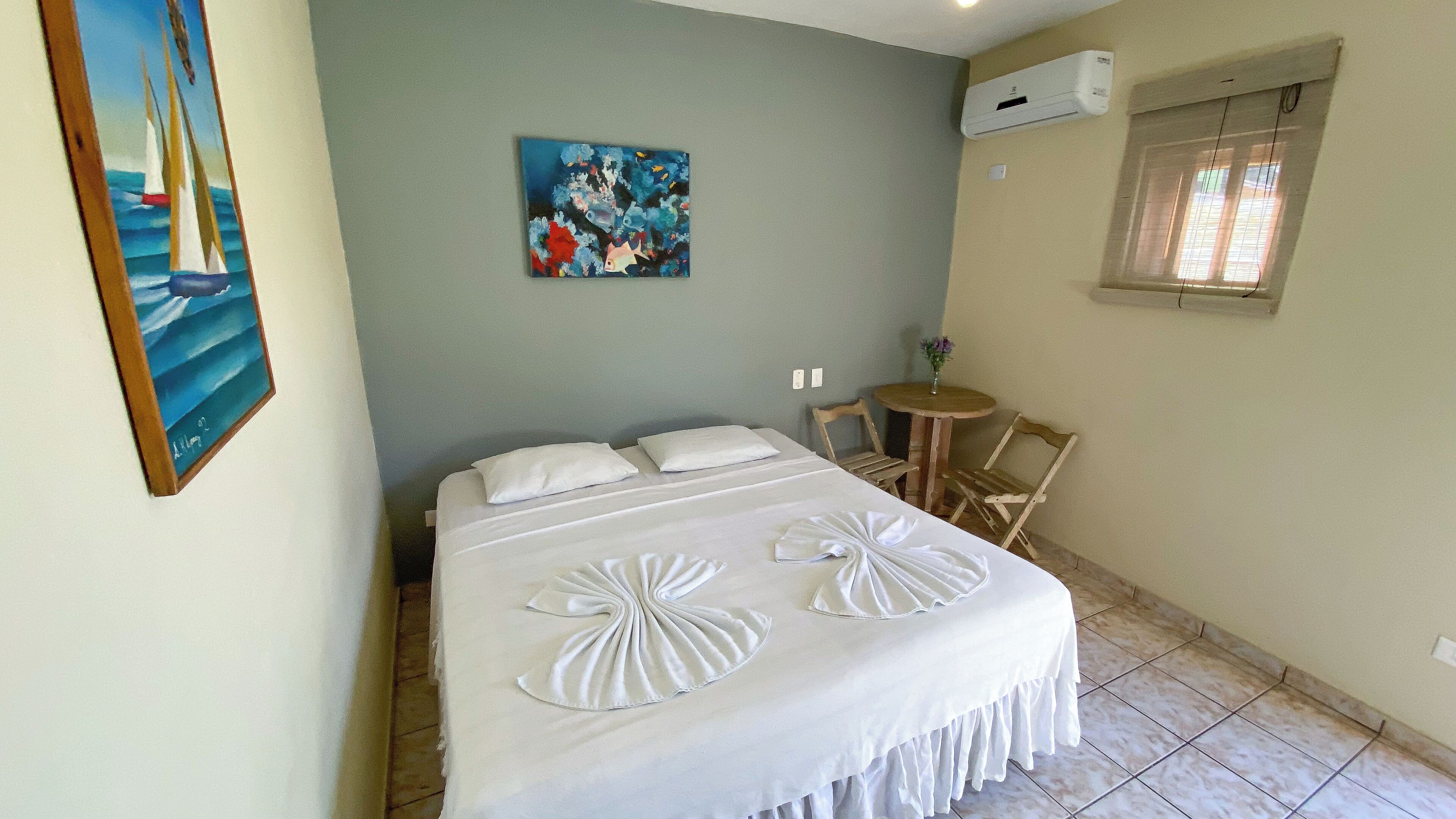 Una cama bien hecha con ropa blanca y almohadones decorativos, una pequeña mesa redonda con un florero y un cuadro de veleros en la pared.