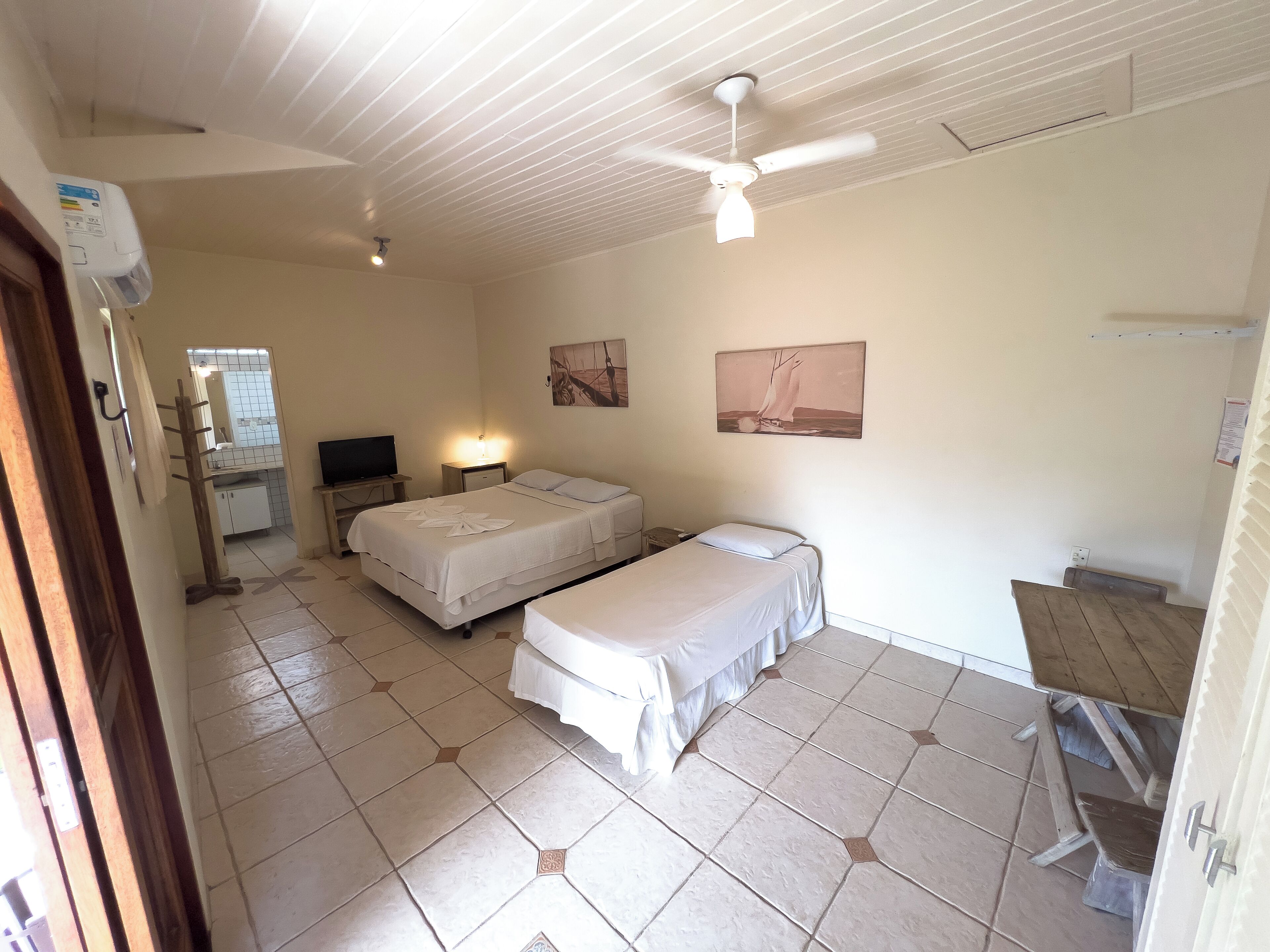 Quarto com duas camas, uma escrivaninha, uma cadeira, ventilador de teto e um quadro na parede.