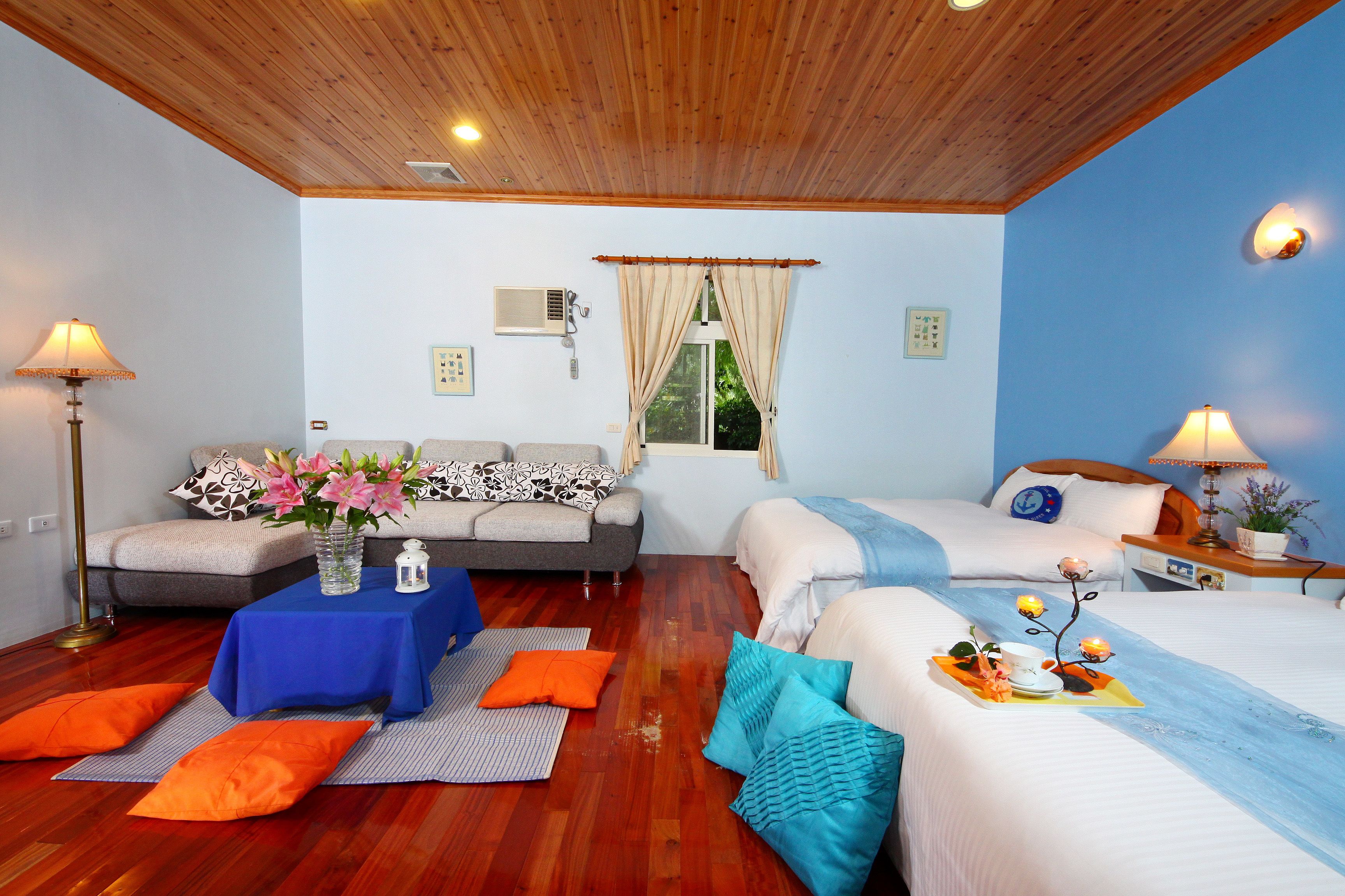 A room with two beds, a sofa, a small table with a vase of flowers, and a window with curtains.