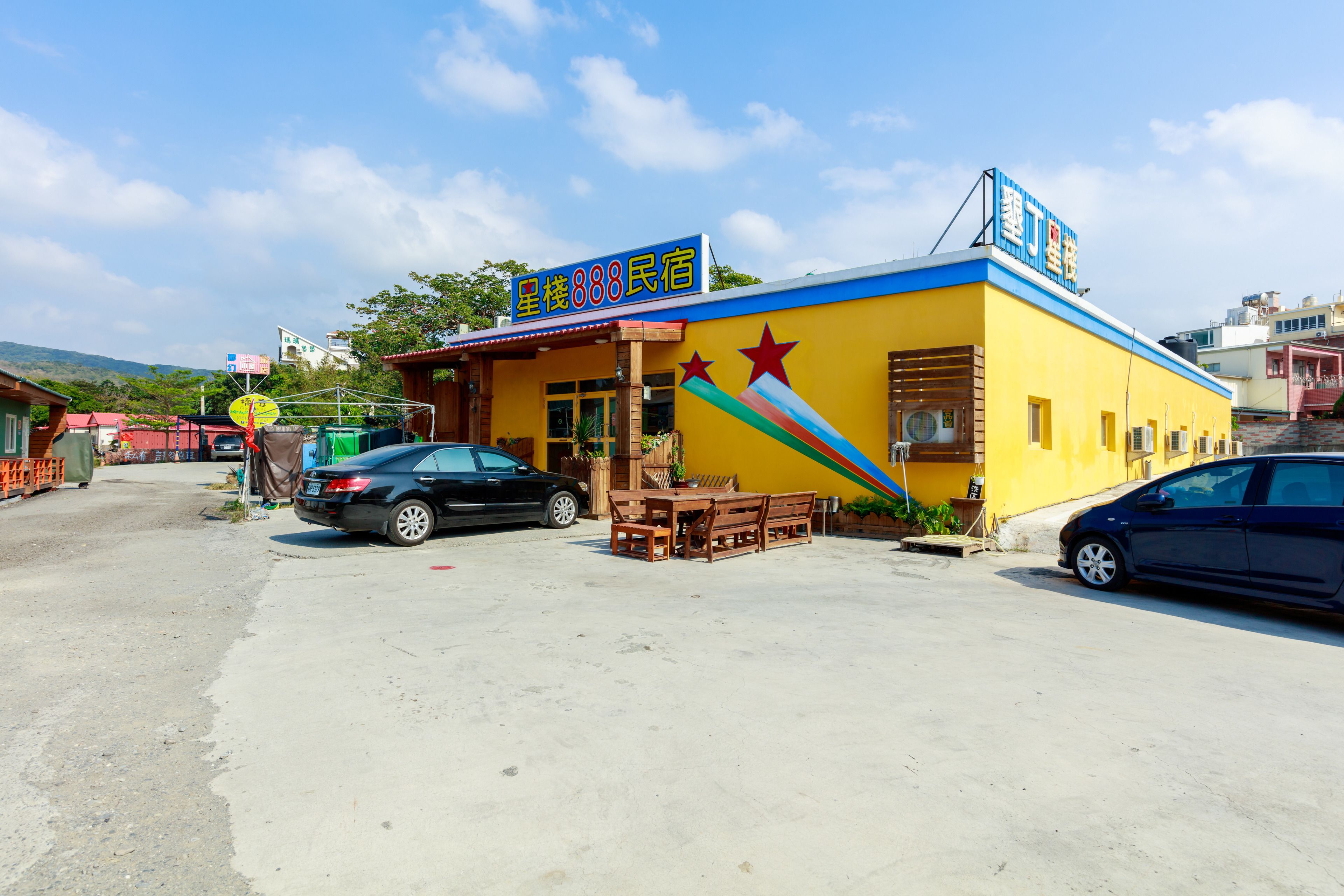 A yellow building with a blue roof and a sign reading "888 Citizen Hotel". There is a wooden table and chairs outside, and two cars parked in front.