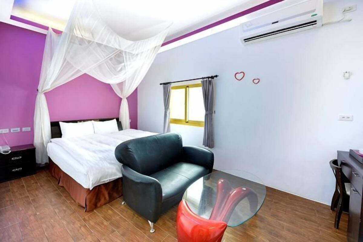 A bedroom with a bed, a chair, a glass table, a window with curtains, and a wall-mounted air conditioner.