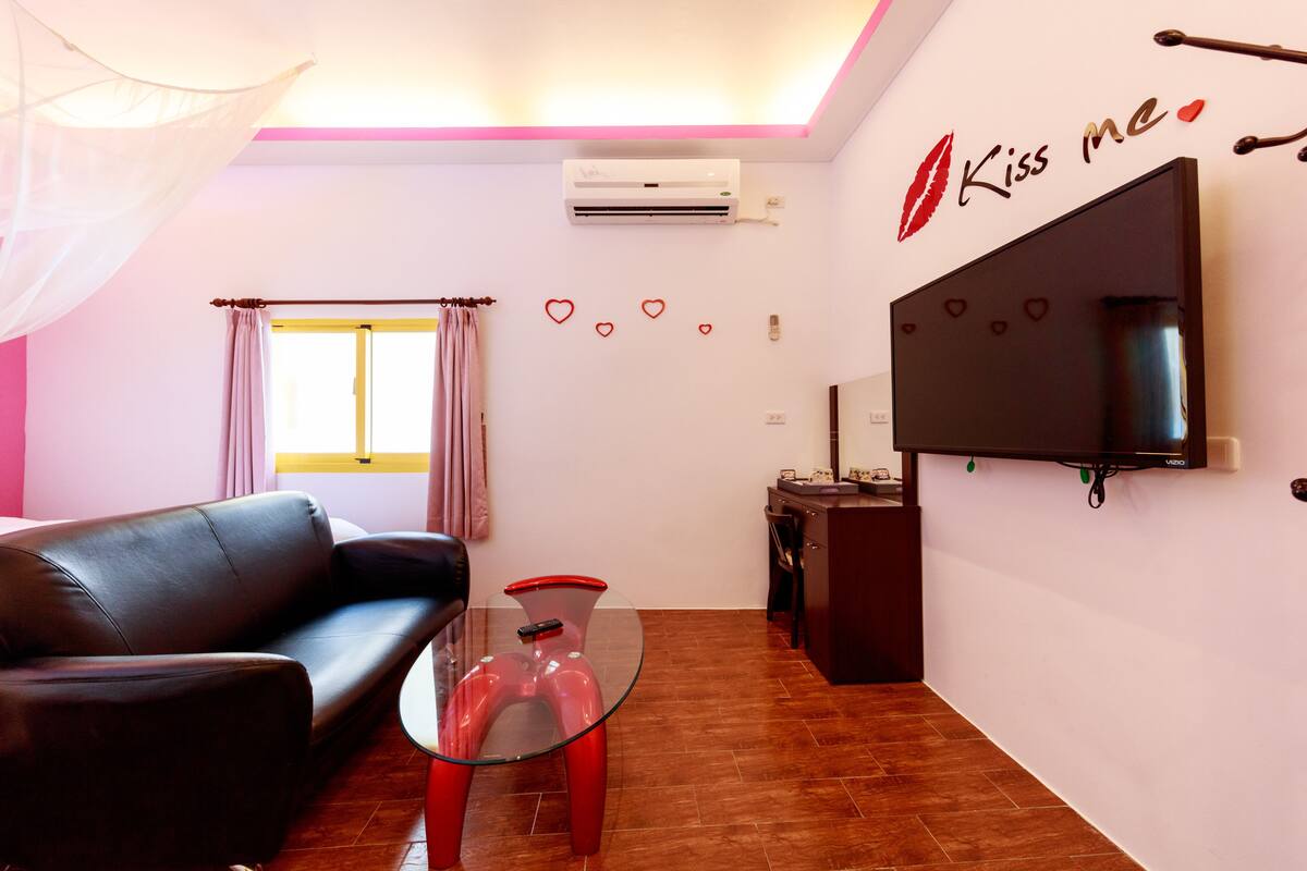 A living room with a black leather sofa, a glass coffee table, a flat-screen TV mounted on the wall, and a window with curtains.