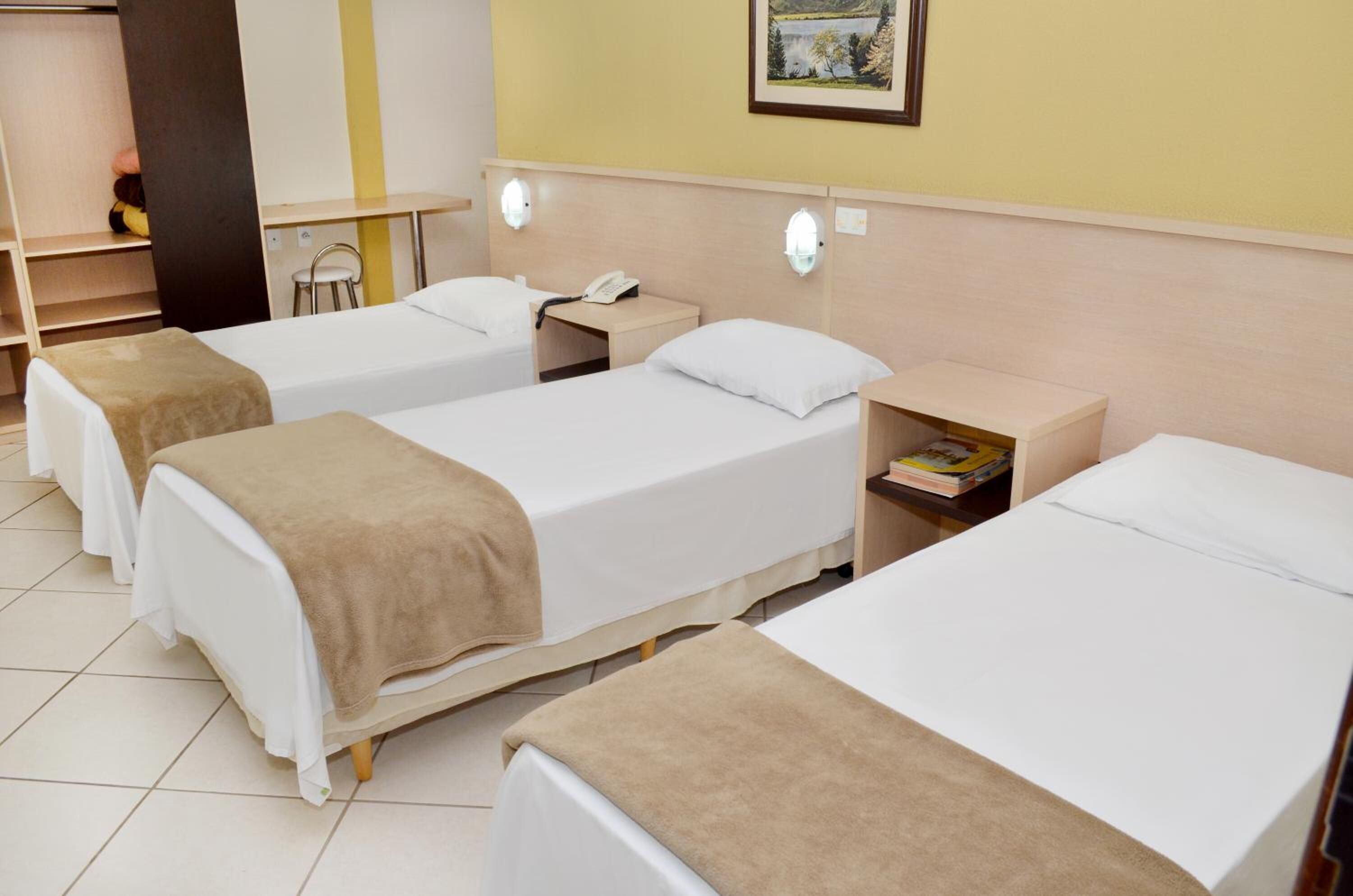 Habitación de hotel con tres camas individuales, cada una con un colchón blanco y una manta marrón. Hay una mesita de noche pequeña con una lámpara y un teléfono. La habitación tiene una pared amarilla y un cuadro enmarcado sobre las camas.