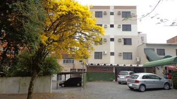 Un estacionamiento con dos coches, un edificio con varias ventanas y unidades de aire acondicionado, y un árbol con flores amarillas.